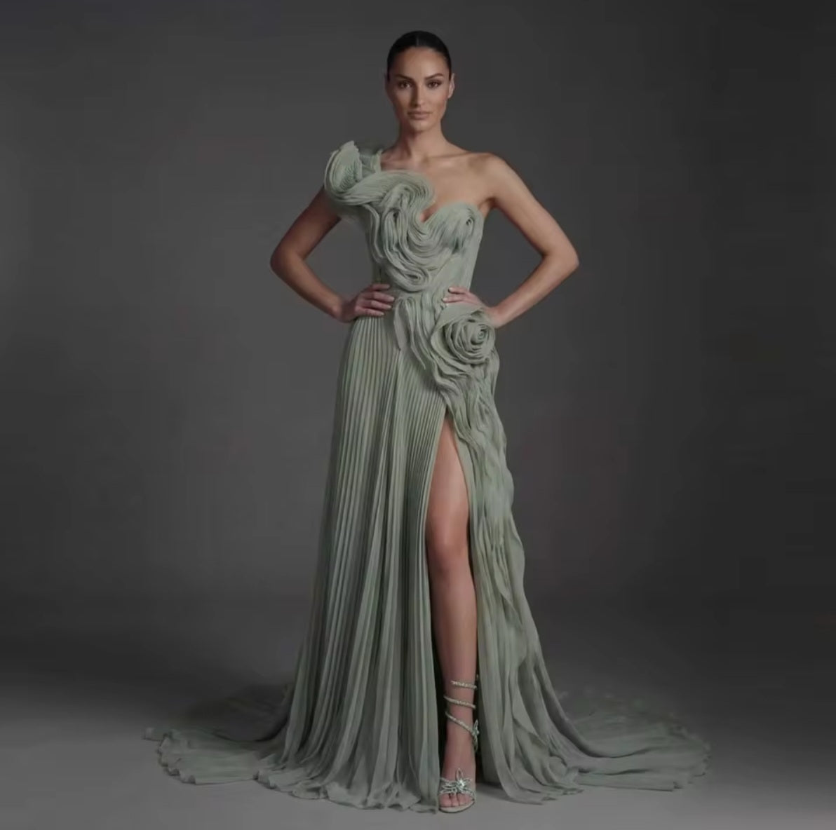 A model wears a floor-length sage green gown with dramatic draped fabric, a one-shoulder design, a high thigh slit, and a long train 